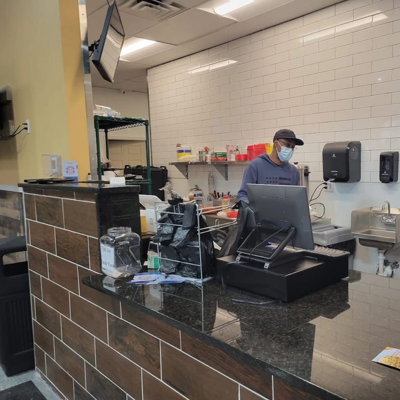 A masked Quruxley Restaurant employee stands behind a counter with a computer and sink.