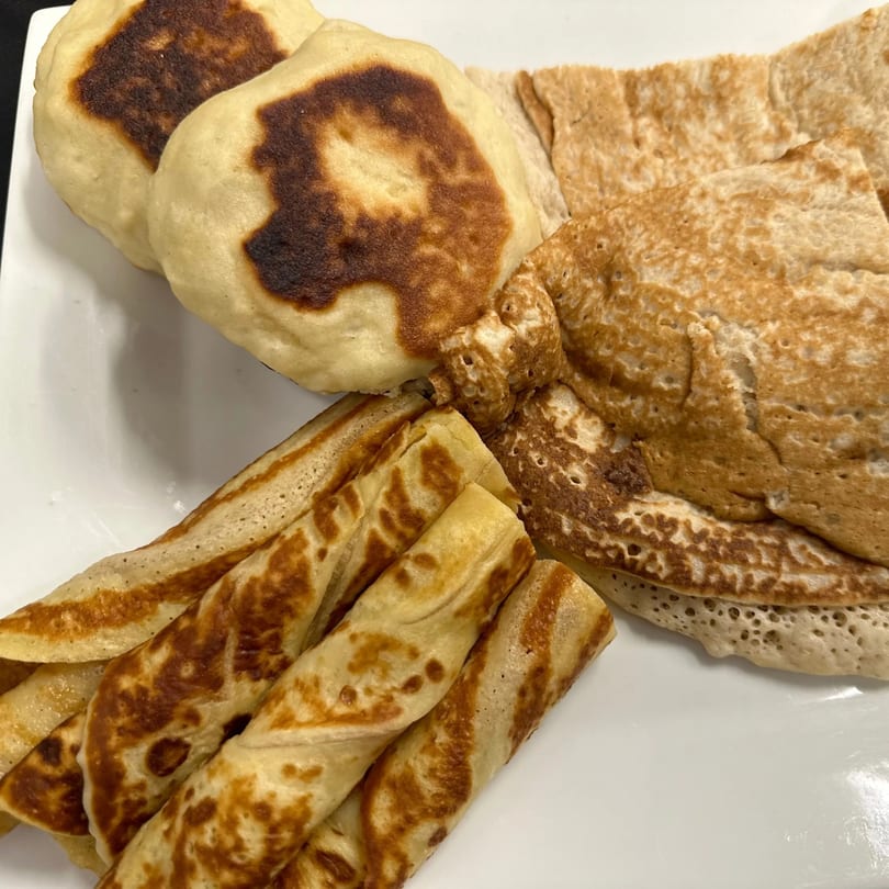 A white plate holds various African flatbreads, including thick, round, browned ones and thin, rolled crepes.