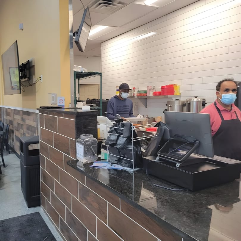Two masked men work behind a counter with a cash register and kitchen supplies at Quruxley Restaurant.