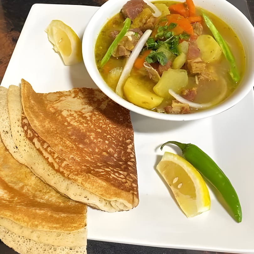A bowl of African stew with meat and vegetables, served with flatbread, lemon, and a green chili.
